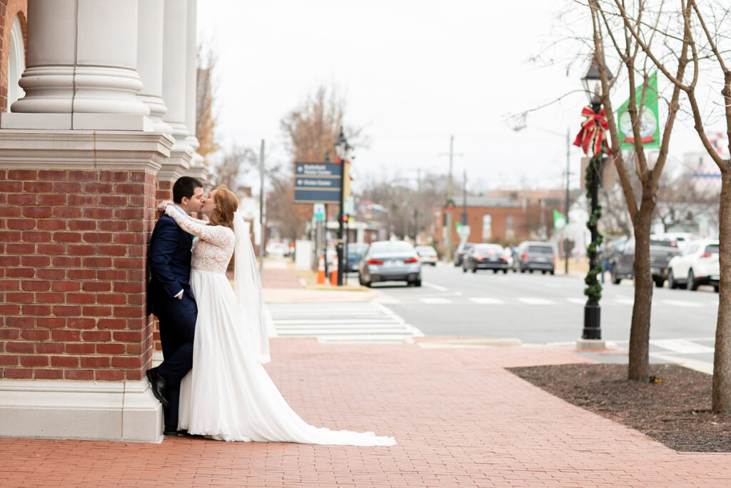 Bride and groom embracing against a brick column downtown, with the bride’s long train spread behind her on the sidewalk.