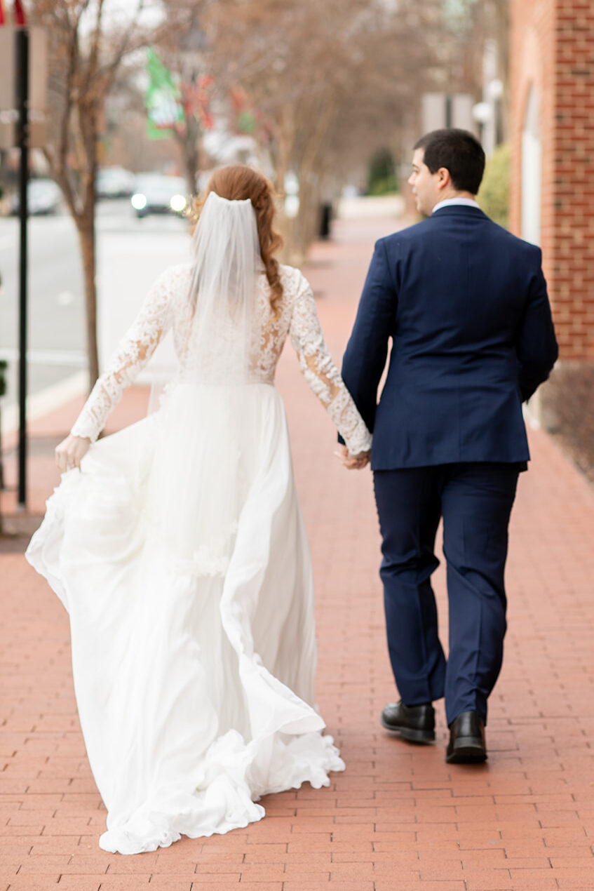 Bride holding up her dress as she walks hand-in-hand with the groom along a brick sidewalk in downtown Fredericksburg.