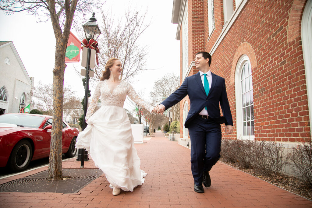 Bride and groom running hand-in-hand down the sidewalk, smiling as they pass a brick building and winter trees.