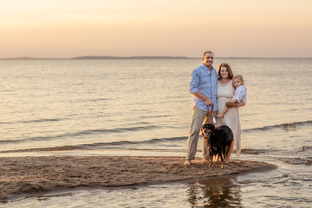 Hope and Tyler standing at the shoreline during their Terrapin Nature Park engagement session, holding their son with their dog nearby as the sun sets over the Chesapeake Bay.