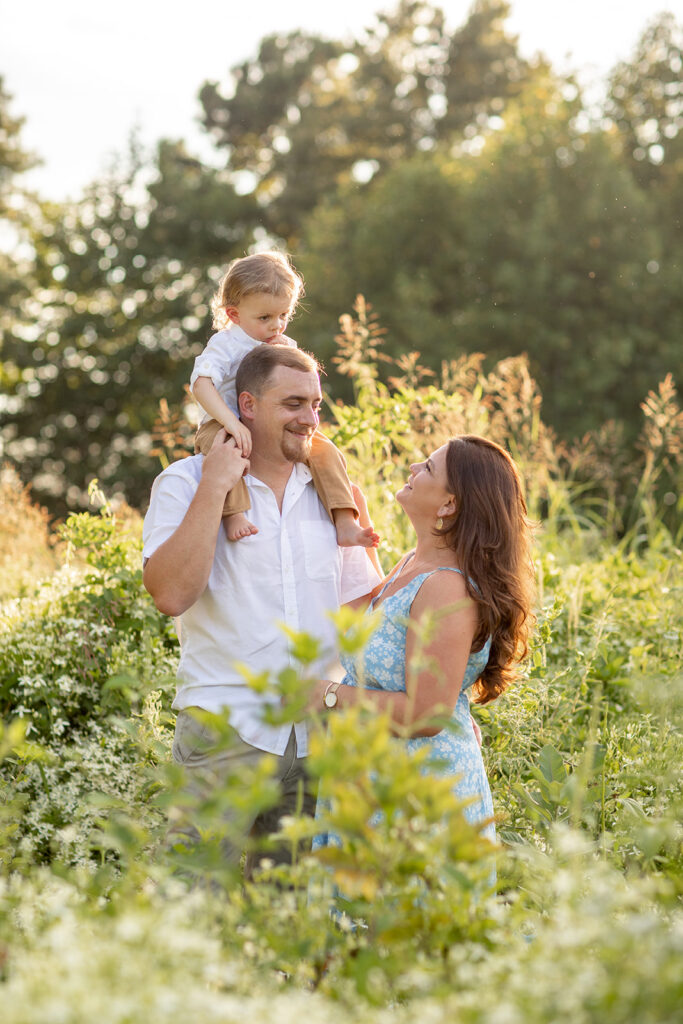 Hope and Tyler laughing while their son sits on Tyler’s shoulders, surrounded by tall grasses at Terrapin Nature Park.