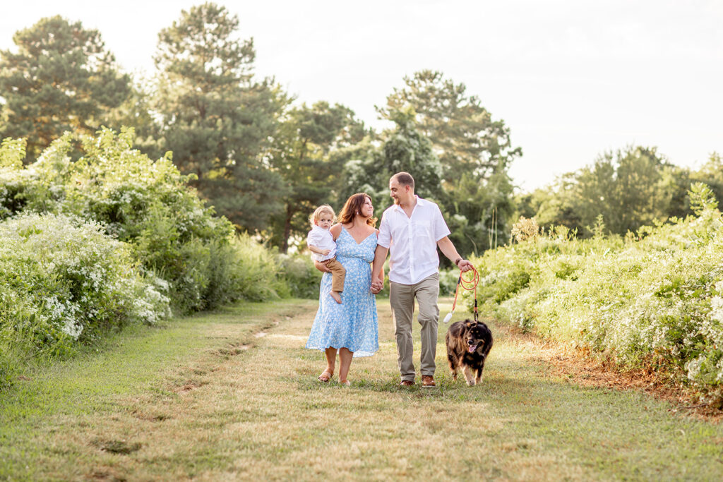 Hope and Tyler walking along a grassy trail at Terrapin Nature Park with their son and dog during their engagement session.