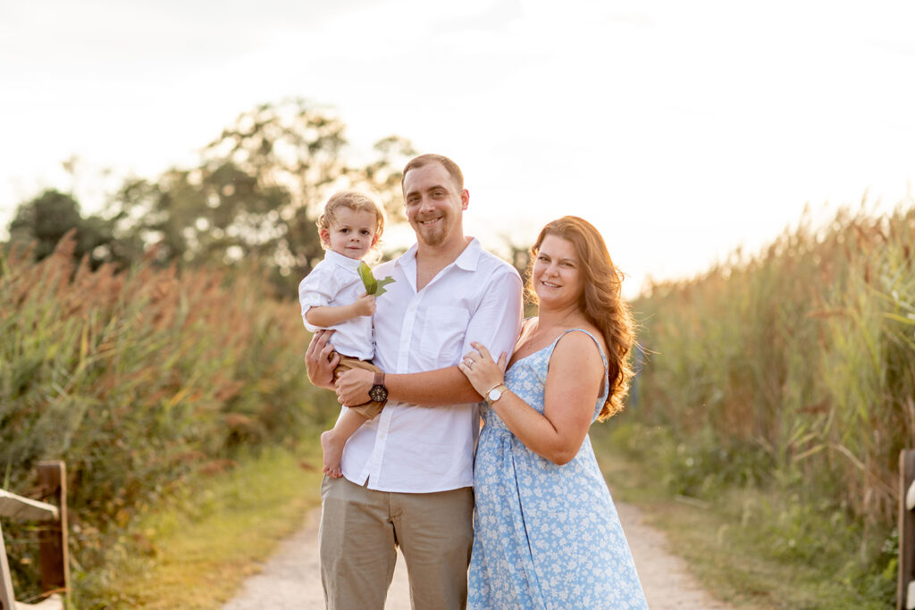 Hope and Tyler smiling at the camera while holding their son on a nature path at Terrapin Nature Park during their engagement session.